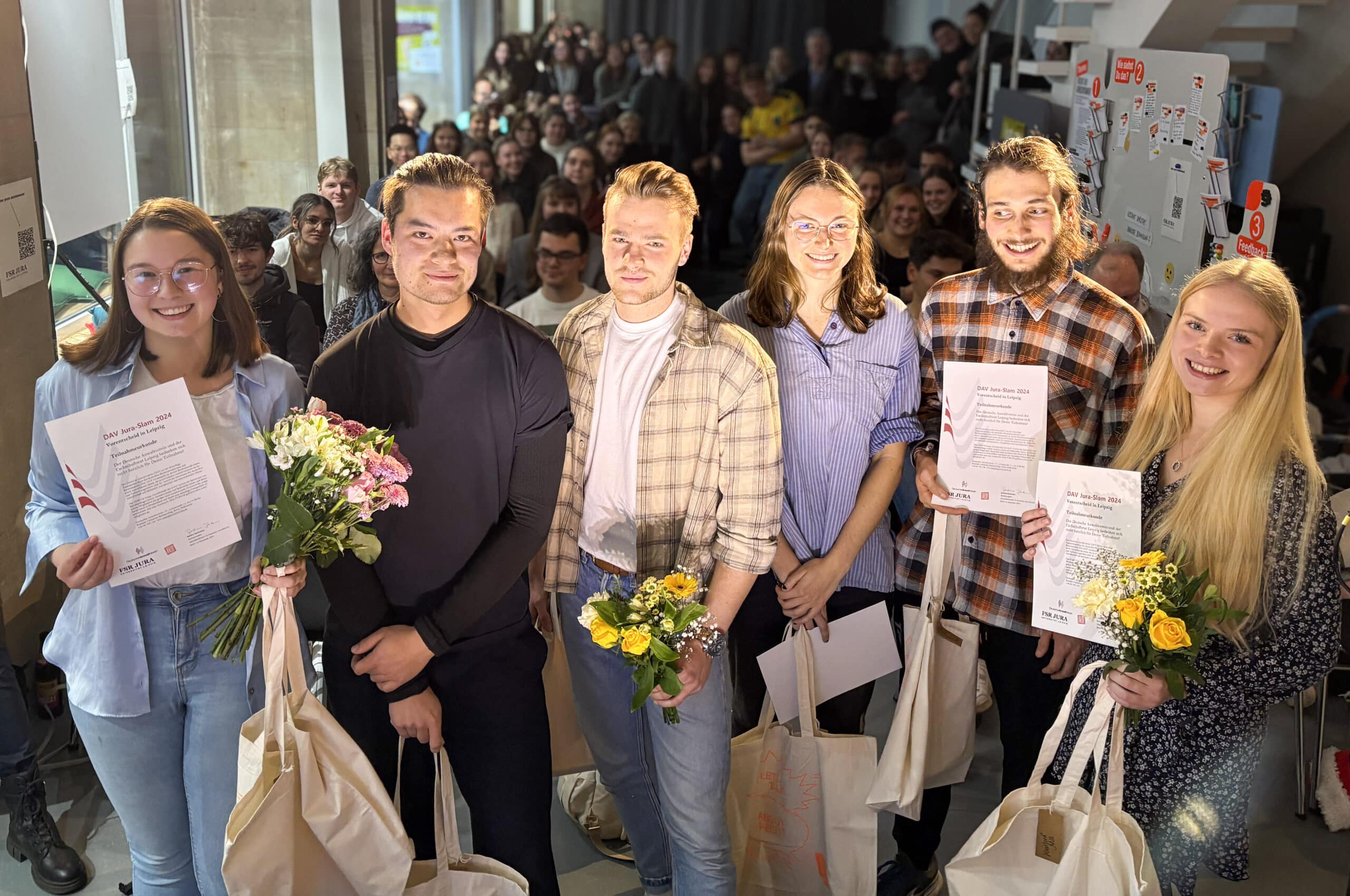 Fünf Studierende stehen mit dem Rücken zum Publikum auf der Bühne und halten Urkunden in der Hand.