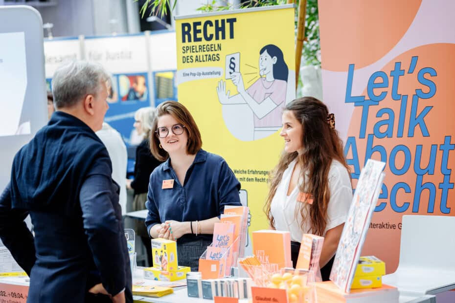 Ein Stand mit orangen Roll-ups im Hintergrund, zwei Frauen und ein Herr im Gespräch.