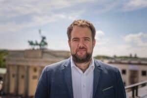 Mann mit Vollbart und dunklen, lockigen haaren vor dem Brandenburger Tor in Berlin.
