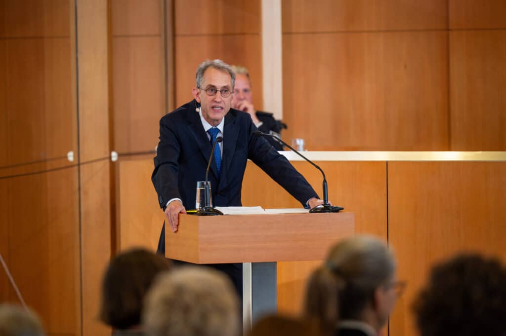 An elderly man with short gray hair and glasses is speaking at a lectern in a wood-paneled courtroom.