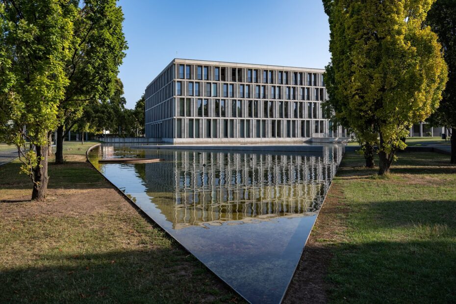 Außenansicht eines modernen Glasbaus in einer Parkanlage mit einer Wasserfläche davor.