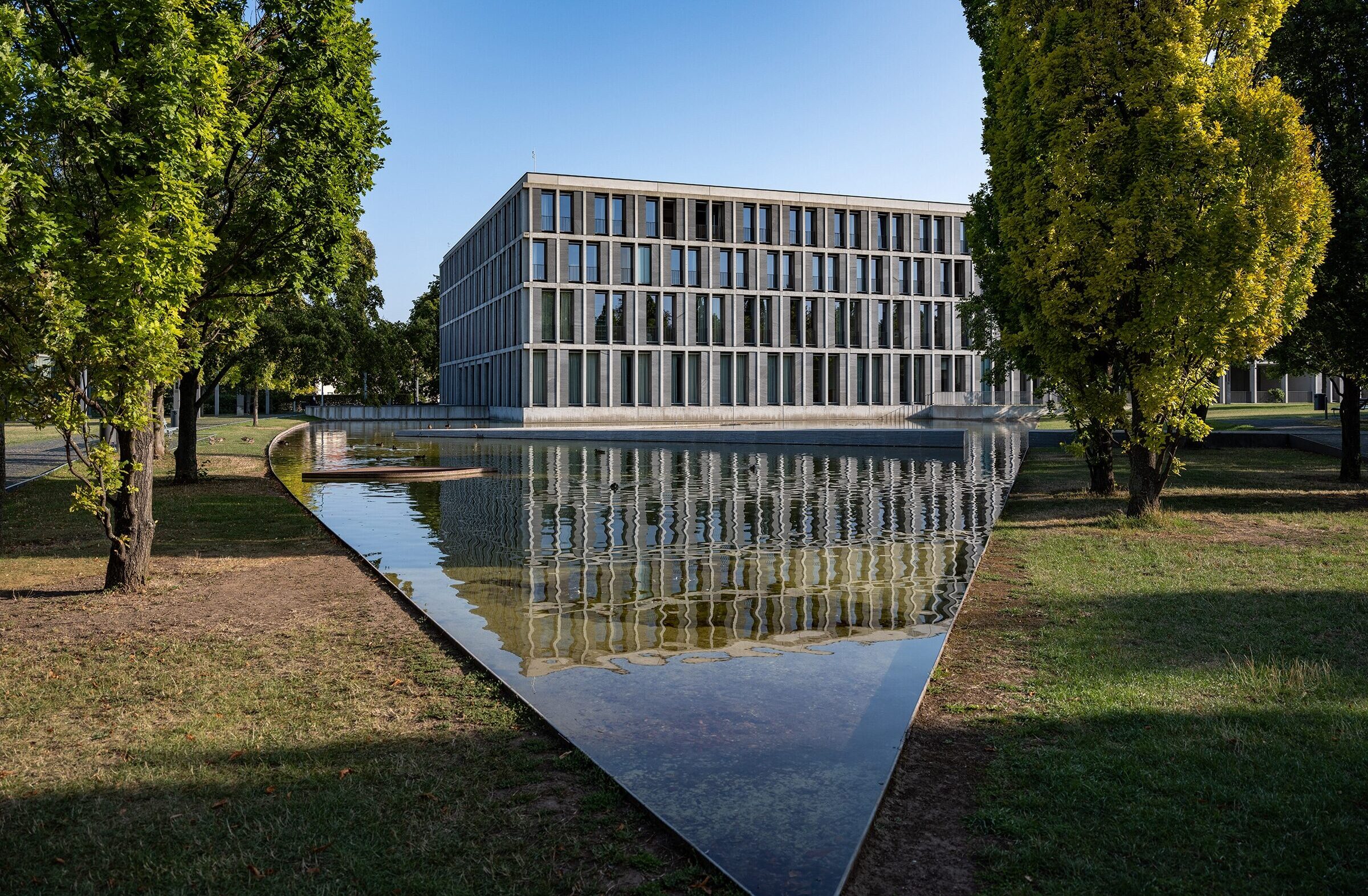 Außenansicht eines modernen Glasbaus in einer Parkanlage mit einer Wasserfläche davor.