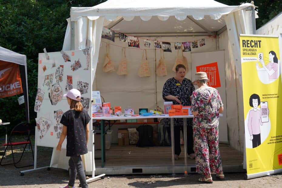 eine Mitarbeiterin der Stiftung Forum Recht steht in einem Pavillon hinter einem Tische mit Merchandise der Stiftung
