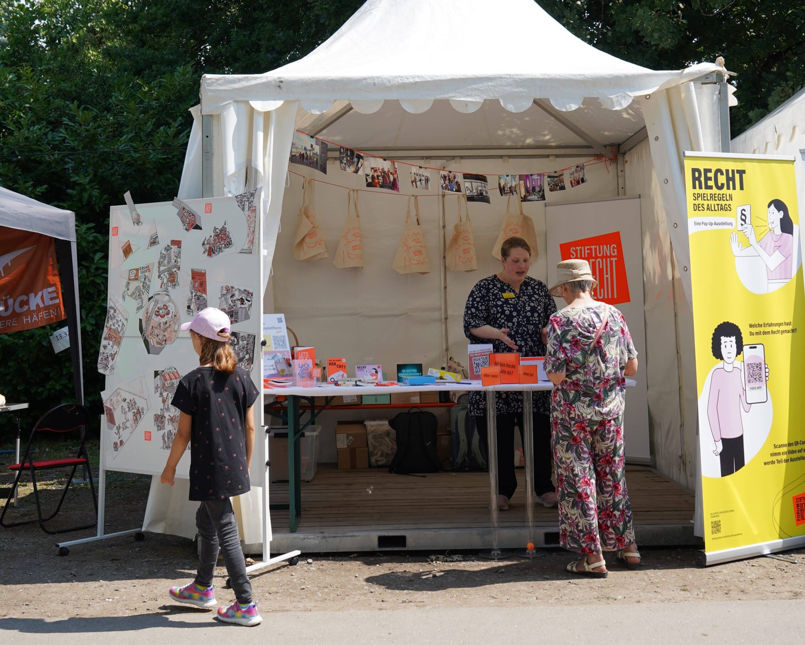 eine Mitarbeiterin der Stiftung Forum Recht steht in einem Pavillon hinter einem Tische mit Merchandise der Stiftung