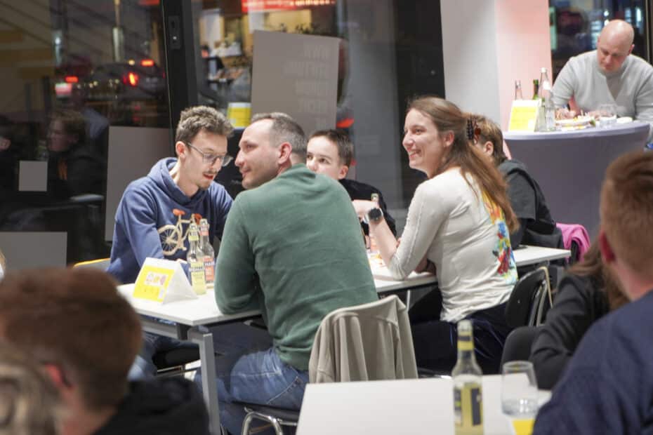 Eine Gruppe von 5 jungen Menschen an einem Tisch blickt lachend zu einer Bühne, die nicht zu sehen ist. Im Hintergrund weitere Menschen.