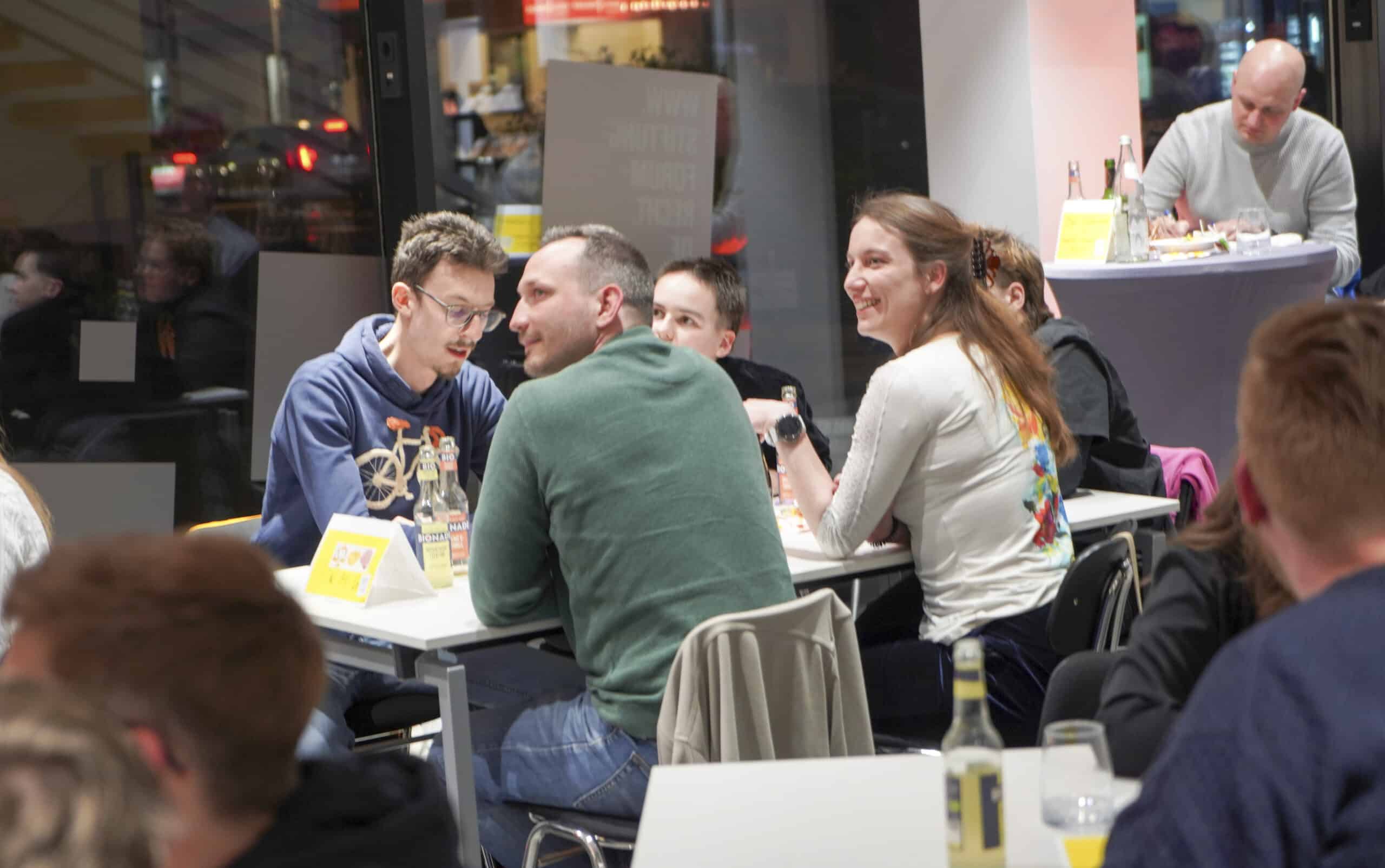 A group of five young people sitting at a table are laughing as they look toward a stage that is out of sight. More people are in the background.